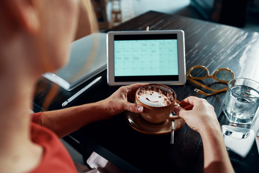 Woman enjoying big cup of cappuccino and checking calendar on tablet computer